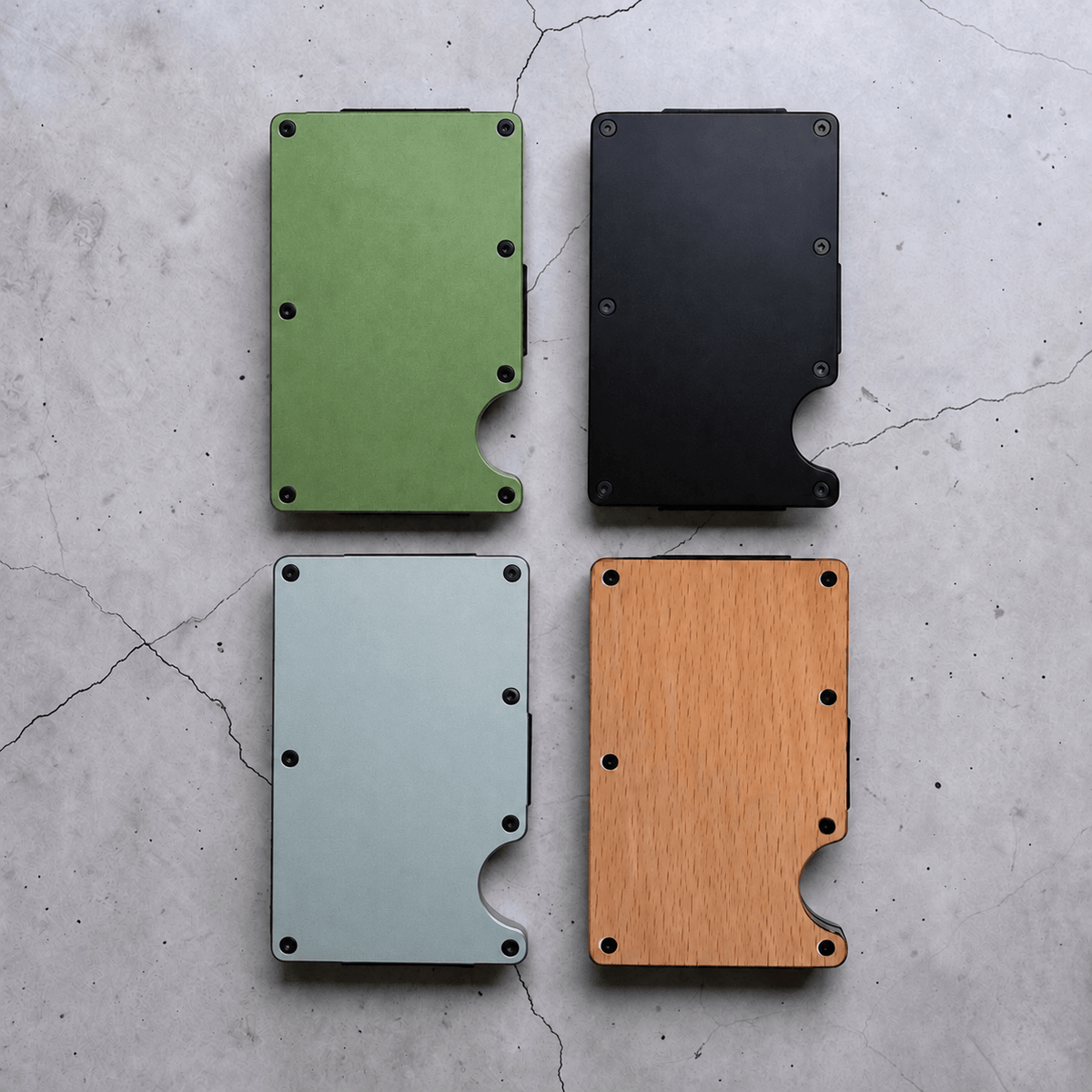 Four slim metal cardholder wallets arranged in a 2x2 grid on a light grey concrete surface. The wallets are shown in four colour options: green, black, light blue, and natural wood grain.