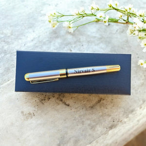 Gold pen with 'Nirvair S' engraving on a blue box, placed on a textured surface with white flowers.