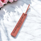 Leather bookmark with engraved quote and tassel beside pink flowers.