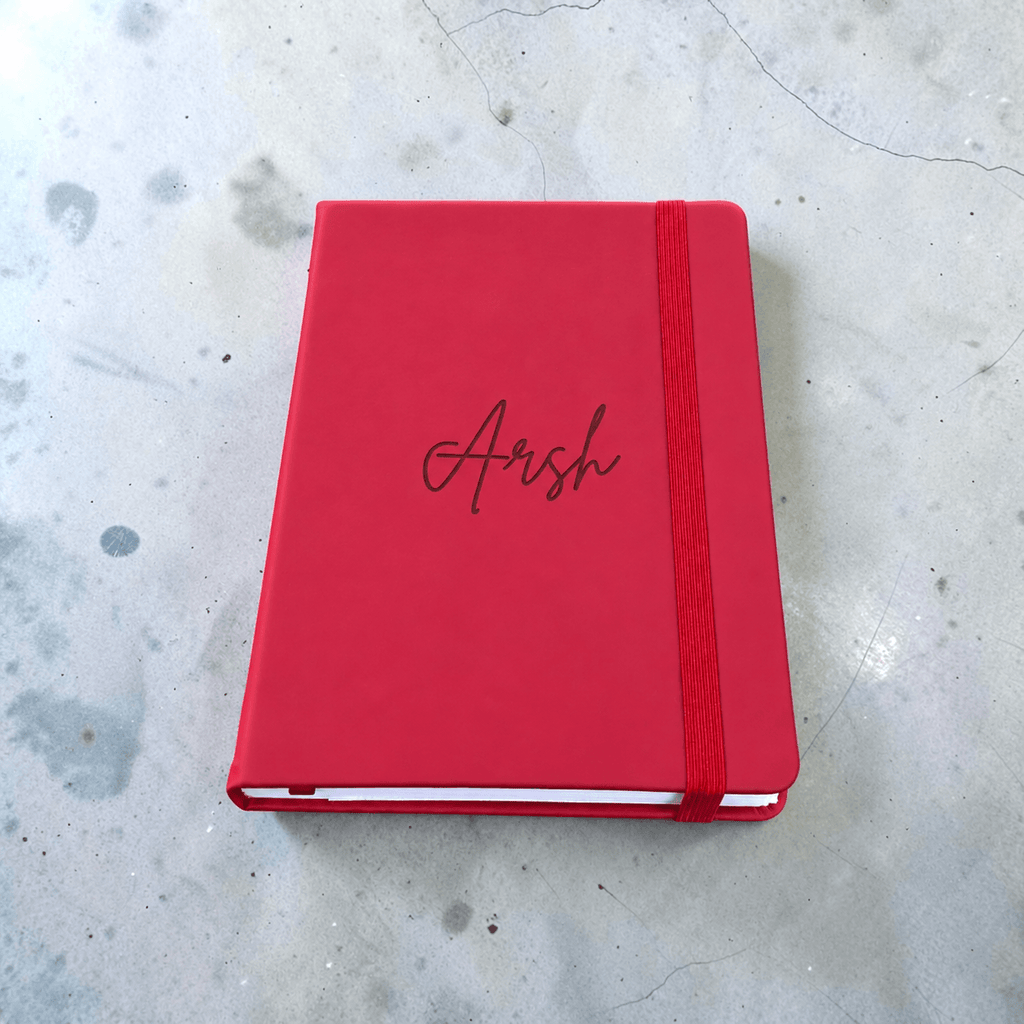 Maroon PU leather journal with Arsh engraved in cursive, showcasing a custom name notebook for writing and journaling, placed on a light gray concrete surface.