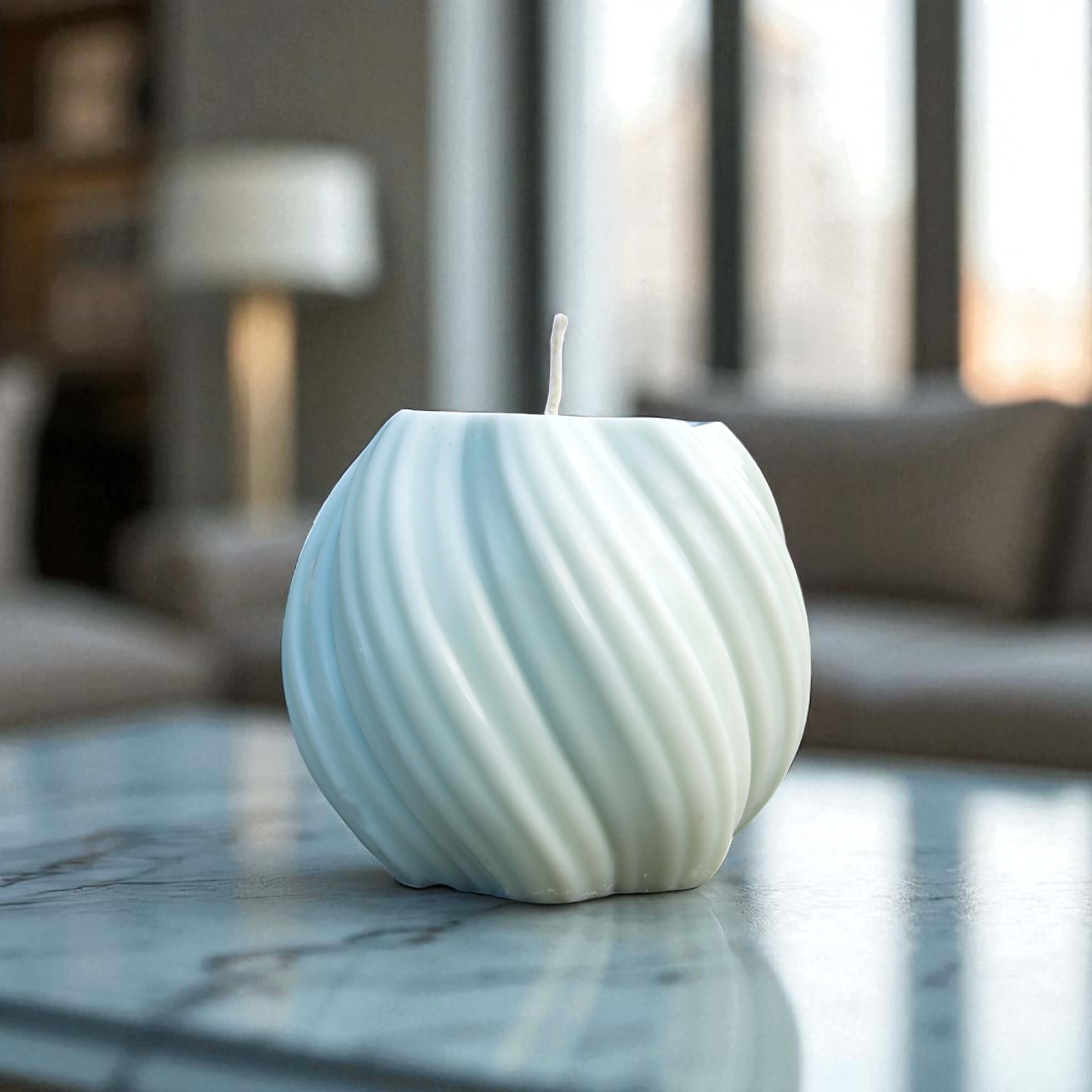 Modern round swirl candle in soft white wax, set on a stone tabletop with natural light from large windows.