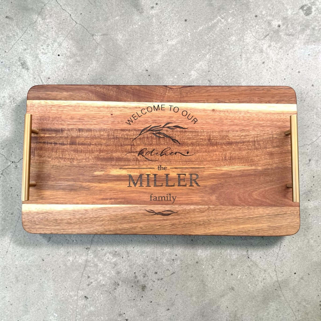 Overhead flat lay of an acacia wood serving tray with gold handles, laser engraved with "Welcome to Our Kitchen" and a personalised family name
