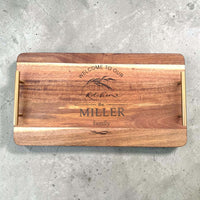 Overhead flat lay of an acacia wood serving tray with gold handles, laser engraved with "Welcome to Our Kitchen" and a personalised family name