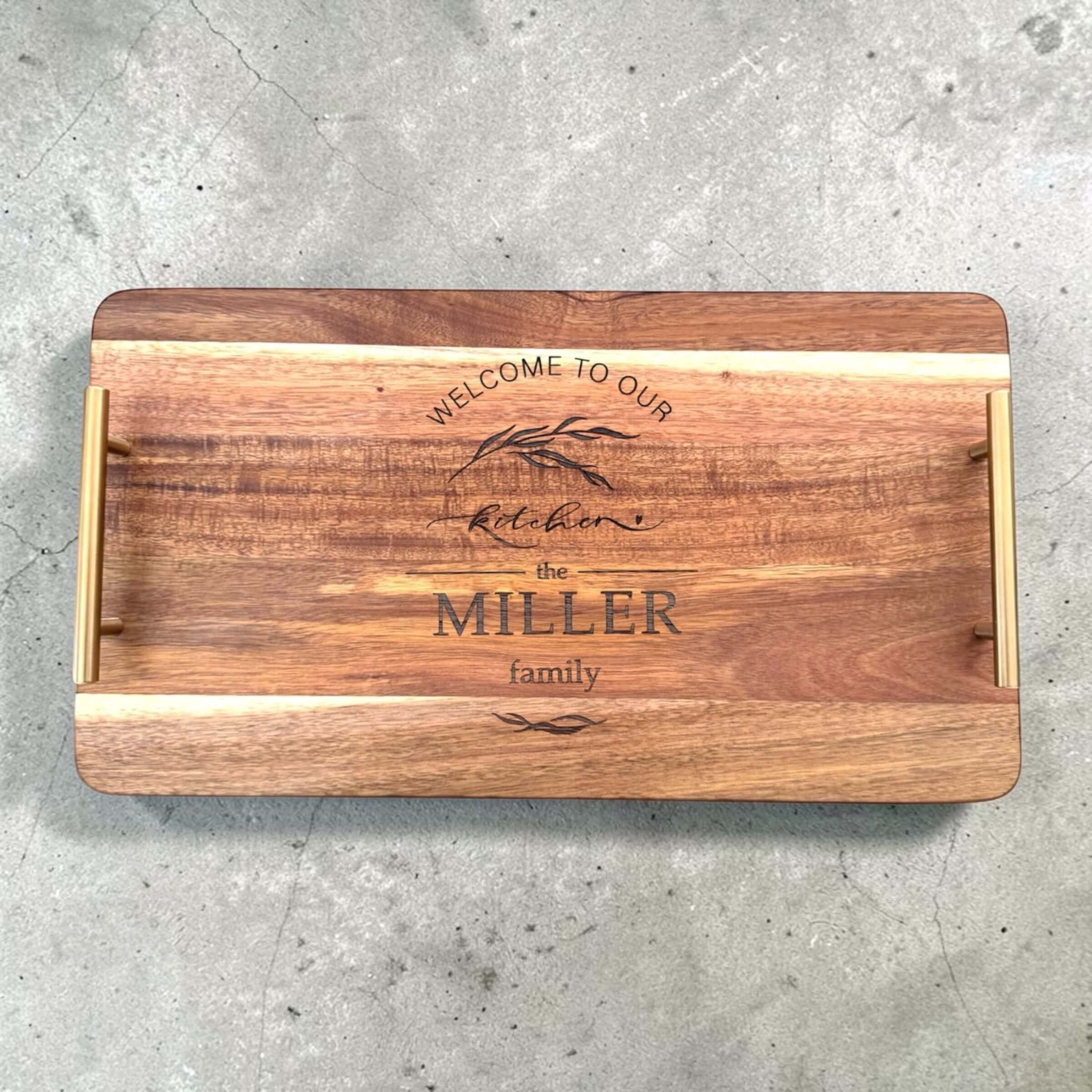 Overhead flat lay of an acacia wood serving tray with gold handles, laser engraved with "Welcome to Our Kitchen" and a personalised family name