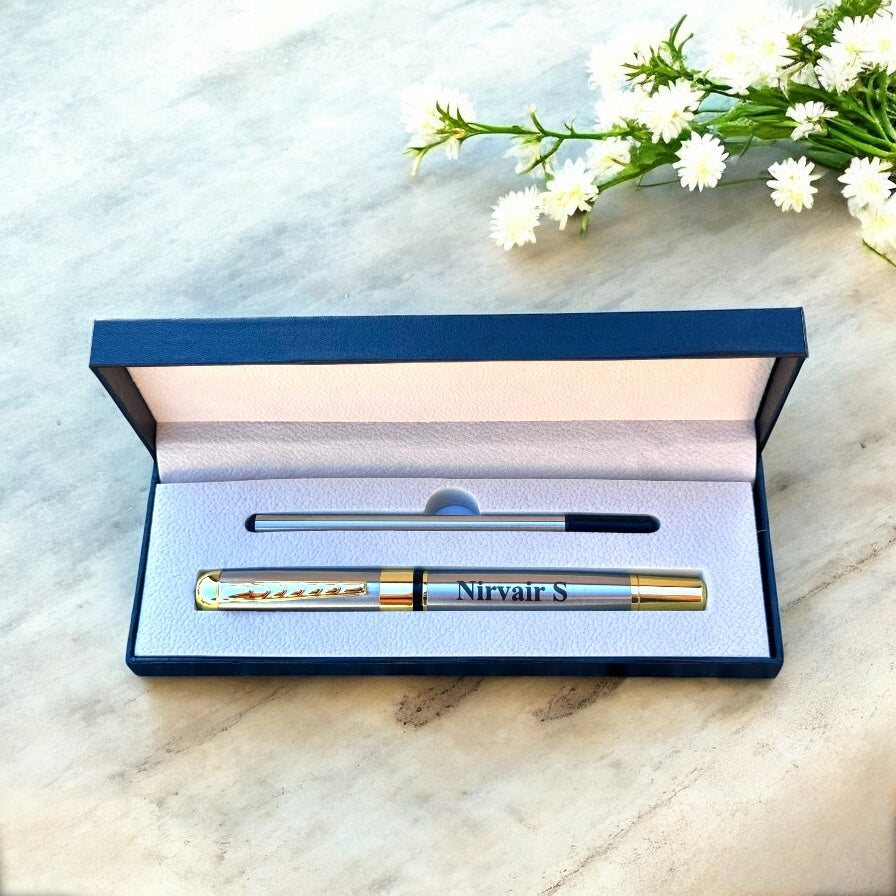 Pen with 'Nirvair S' engraving in an open blue box on a marble surface with white flowers.