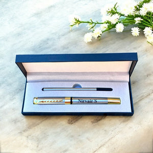 Pen with 'Nirvair S' engraving in an open blue box on a marble surface with white flowers.