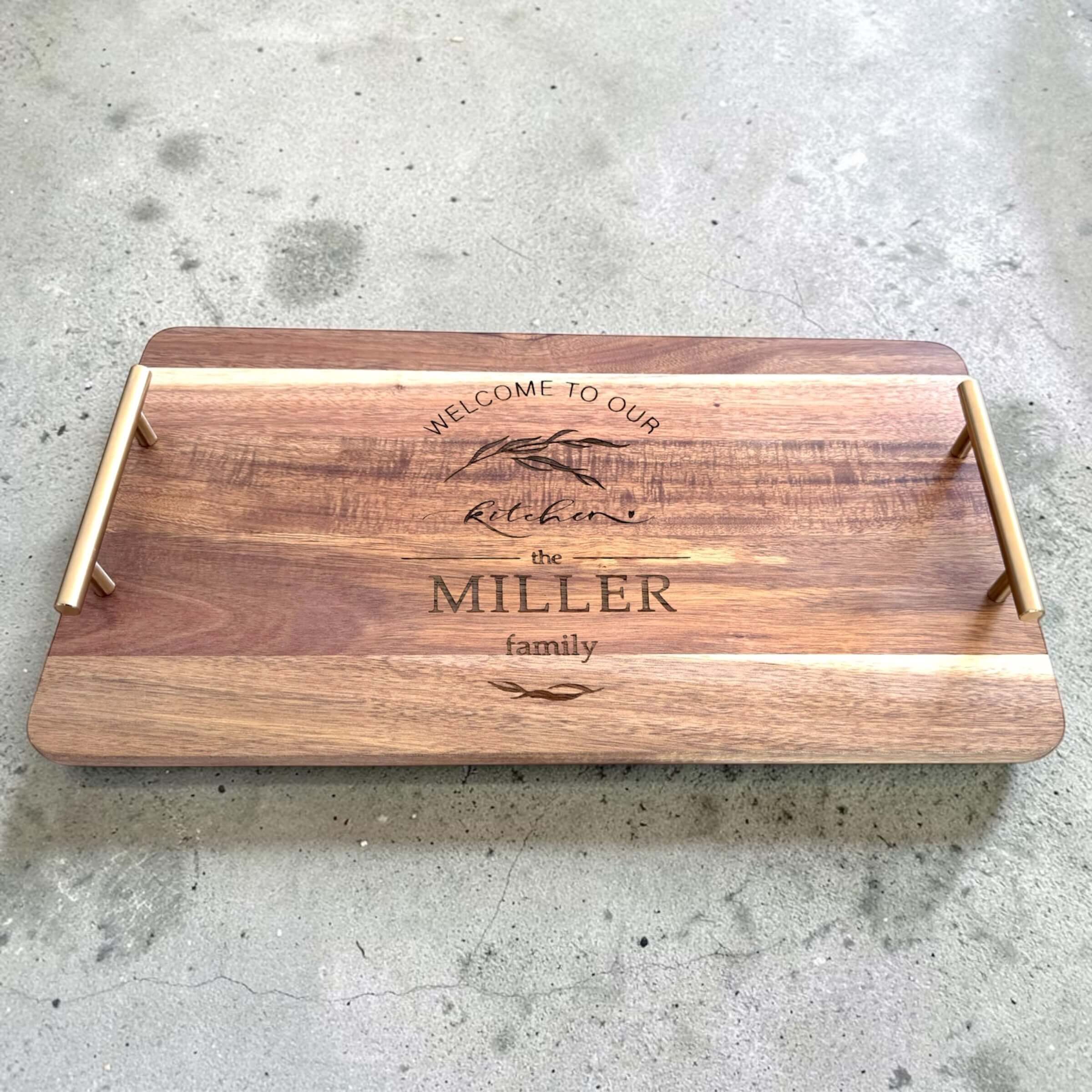 Personalised acacia wood serving platter engraved with a botanical kitchen design and custom family name, photographed from a slight elevation