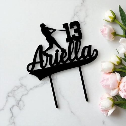 Personalised baseball cake topper featuring a batting figure, age number, and flowing name cutout, presented with soft pink and white flowers.