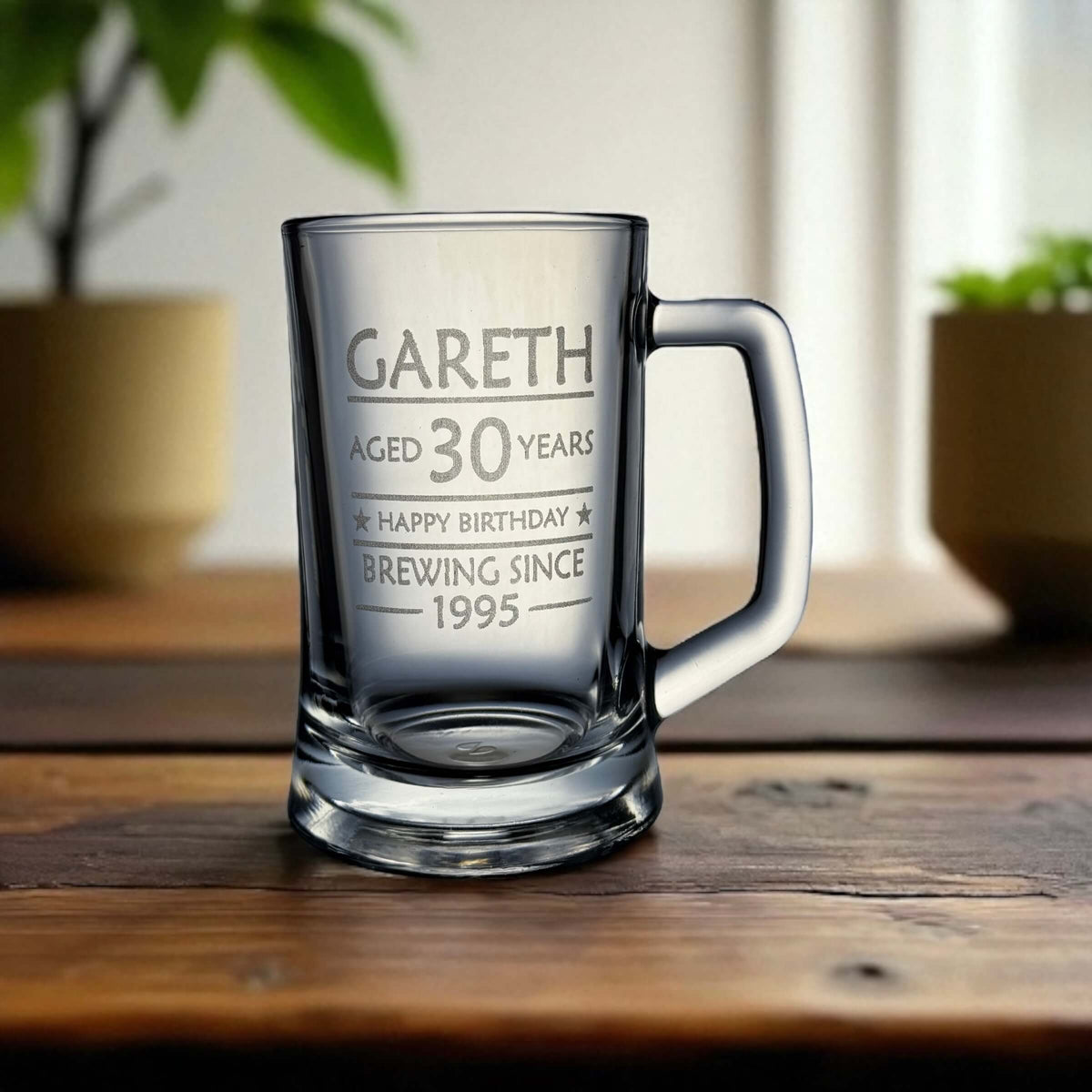 Personalised engraved beer mug with handle featuring the name Gareth and text 'Aged 30 years, Happy Birthday, Brewing Since 1995' placed on a rustic wooden table with blurred potted plants in the background