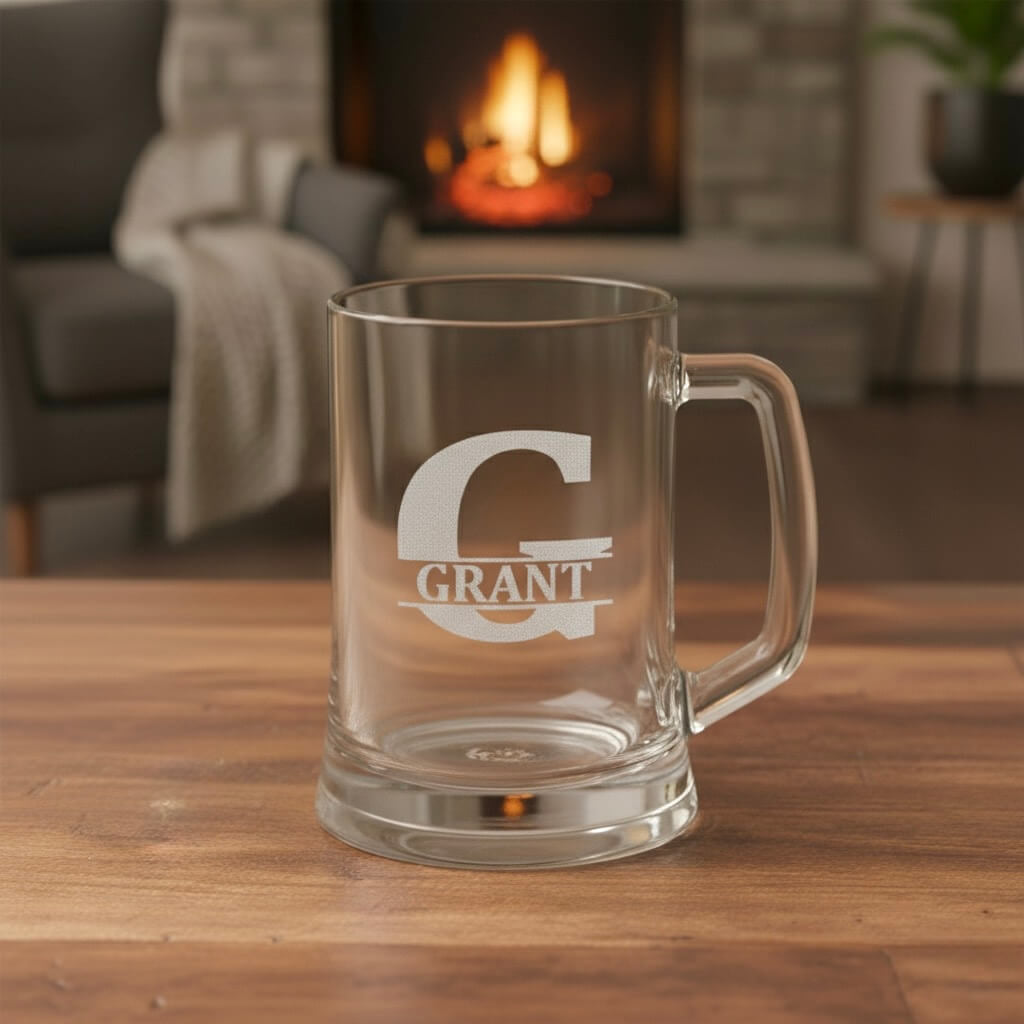 Personalised glass beer mug engraved with large letter G and name Grant, warm firelight background