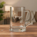 Personalised glass beer mug engraved with the name George, displayed on wooden table in warm home setting