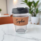 Personalised glass coffee cup with cork sleeve engraved “Sophie” and black lid on white marble table