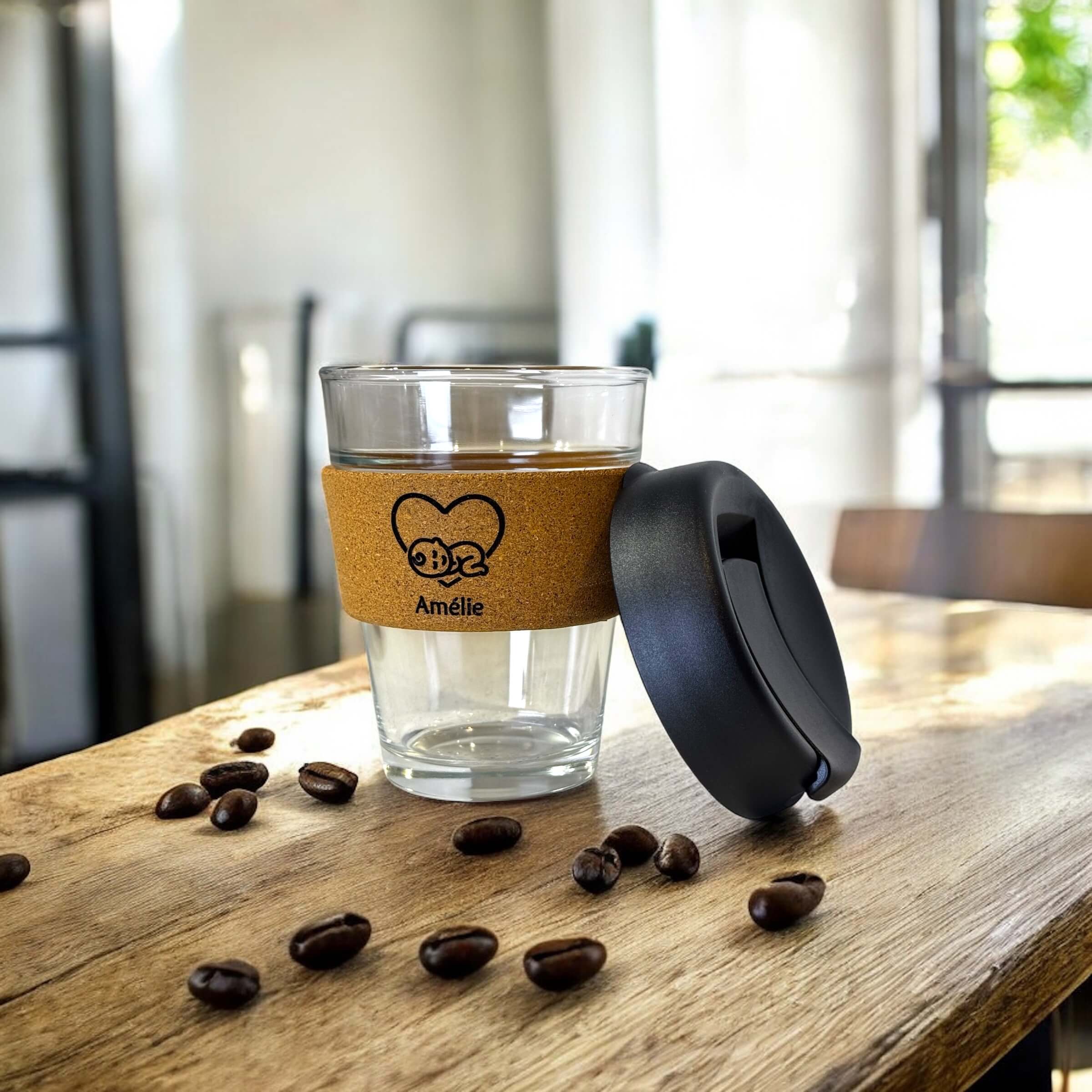 Personalised reusable coffee cup with cork sleeve and the name Amélie, placed on a wooden surface next to scattered roasted coffee beans with the lid resting beside it in a sunlit café environment