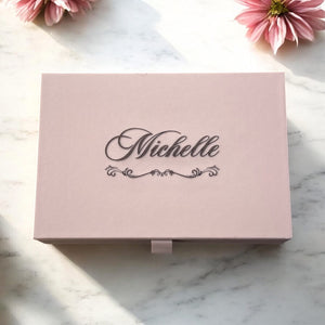 Personalised taupe jewellery box with engraved name on lid in elegant script, surrounded by soft pink flowers.