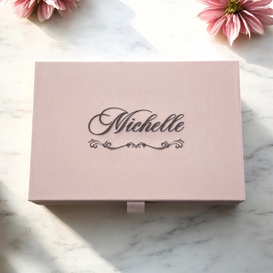 Personalised taupe jewellery box with engraved name on lid in elegant script, surrounded by soft pink flowers.