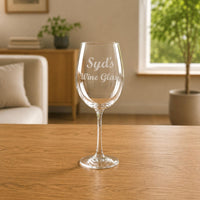 Personalised wine glass engraved with the name Syd in a script font, sitting on a wooden table in a cosy living room setting