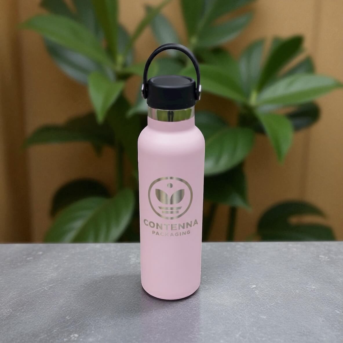 Pink engraved stainless steel drink bottle shown upright on dark stone surface with plants in background.