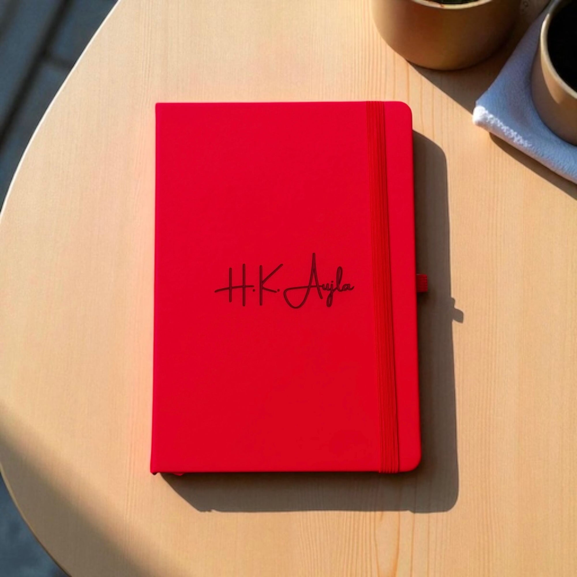 Red notebook with engraved name 'HK Aujla' on a wooden surface