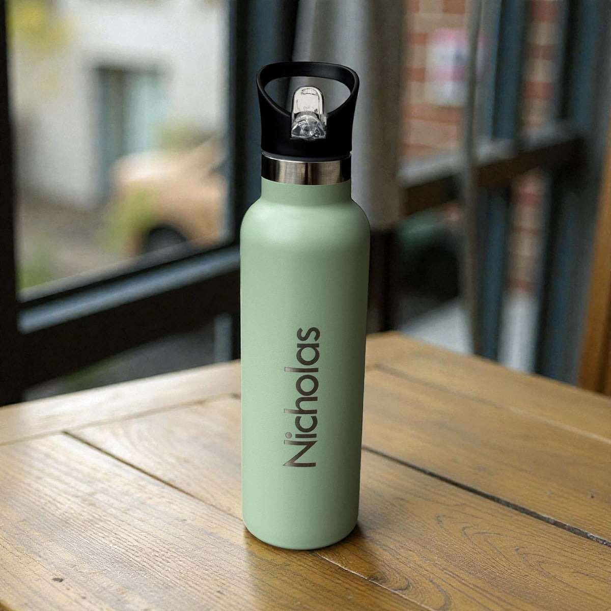 Sage personalised drink bottle with vertical name engraving and a black flip valve lid on a wooden dining table.