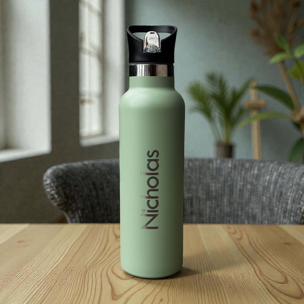 Sage stainless steel water bottle engraved with Nicholas, with a flip valve lid, on a wooden table beside a window.