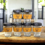 Set of six personalised reusable coffee cups with cork bands, black lids and names Saradha, Christine, Carol, Michelle, Emma and Amélie, displayed on a wooden table in a modern café setting