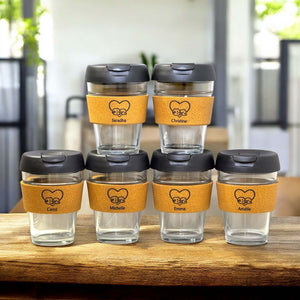 Set of six personalised reusable coffee cups with cork bands, black lids and names Saradha, Christine, Carol, Michelle, Emma and Amélie, displayed on a wooden table in a modern café setting