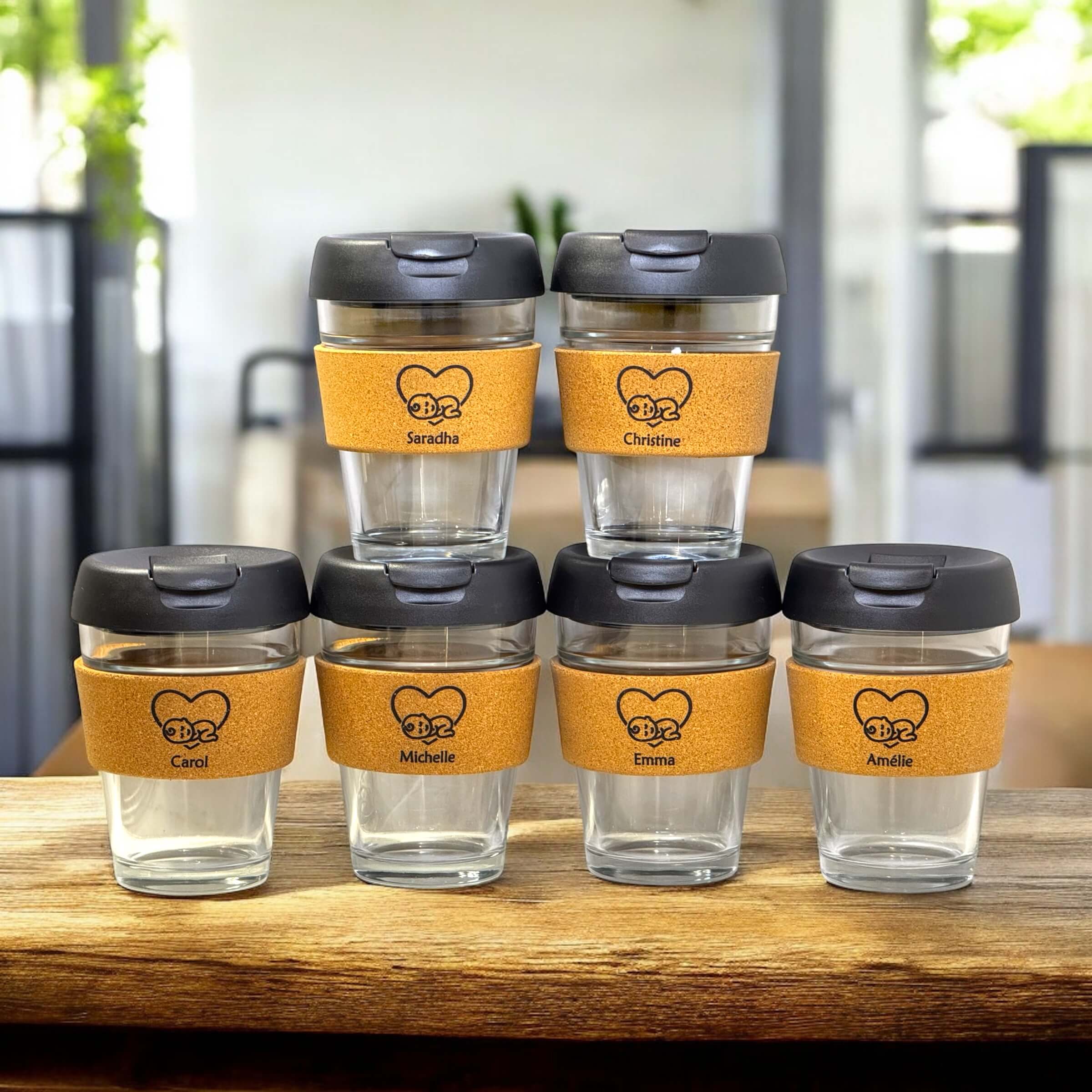 Set of six personalised reusable coffee cups with cork bands, black lids and names Saradha, Christine, Carol, Michelle, Emma and Amélie, displayed on a wooden table in a modern café setting