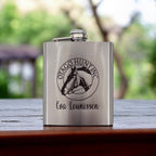 Silver stainless steel hip flask engraved with company logo and name, styled outdoors on a wooden table.