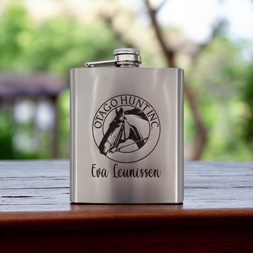 Silver stainless steel hip flask engraved with company logo and name, styled outdoors on a wooden table.