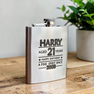 Stainless steel flask with engraved name, age, happy birthday and year born on a wooden surface with a plant in the background