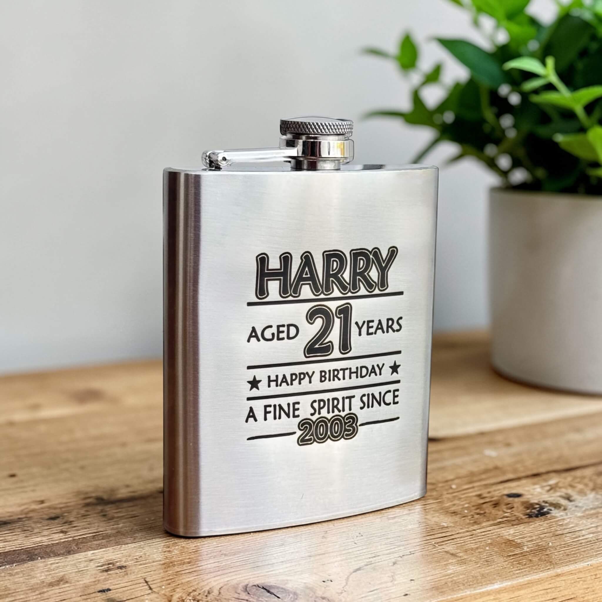 Stainless steel flask with engraved name, age, happy birthday and year born on a wooden surface with a plant in the background