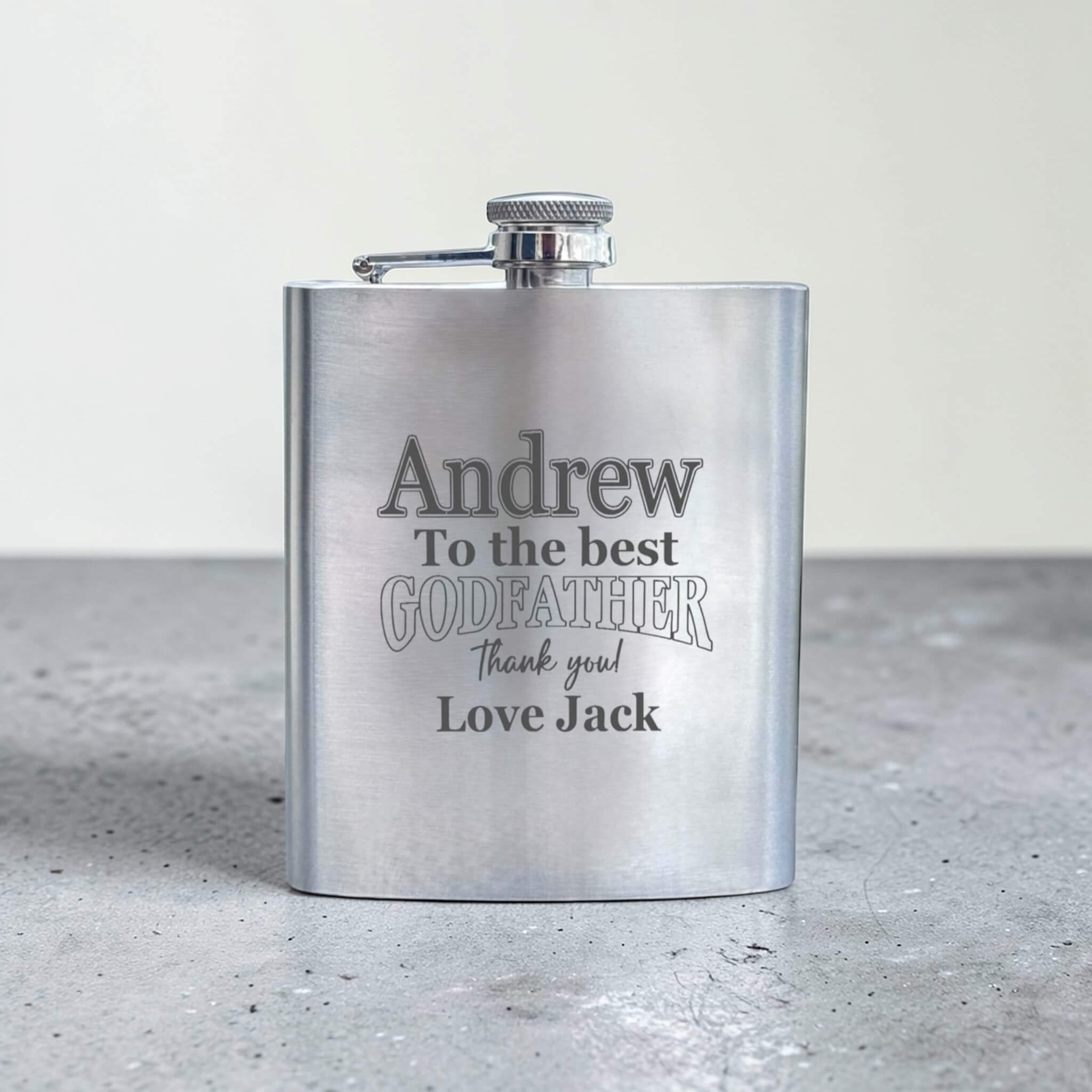 Stainless steel flask with engraved text for godfather on a gray surface