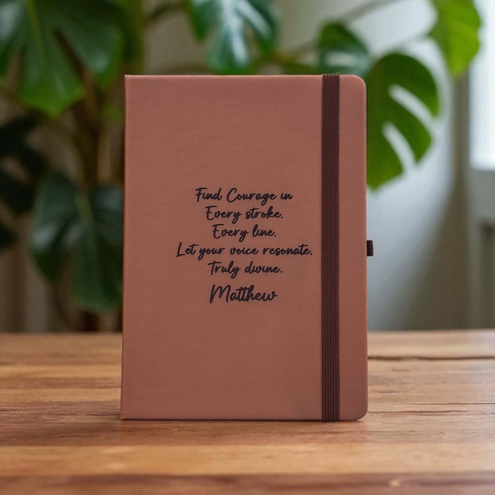 Tan notebook with personalised motivational quote on a wooden surface with plants in the background