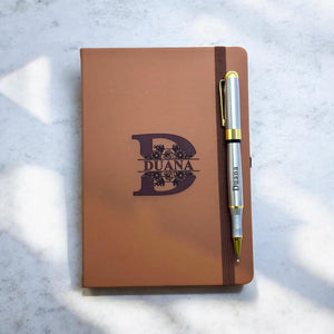 Tan personalised notebook engraved with floral split initial and name, paired with a custom metal pen clipped to the elastic band