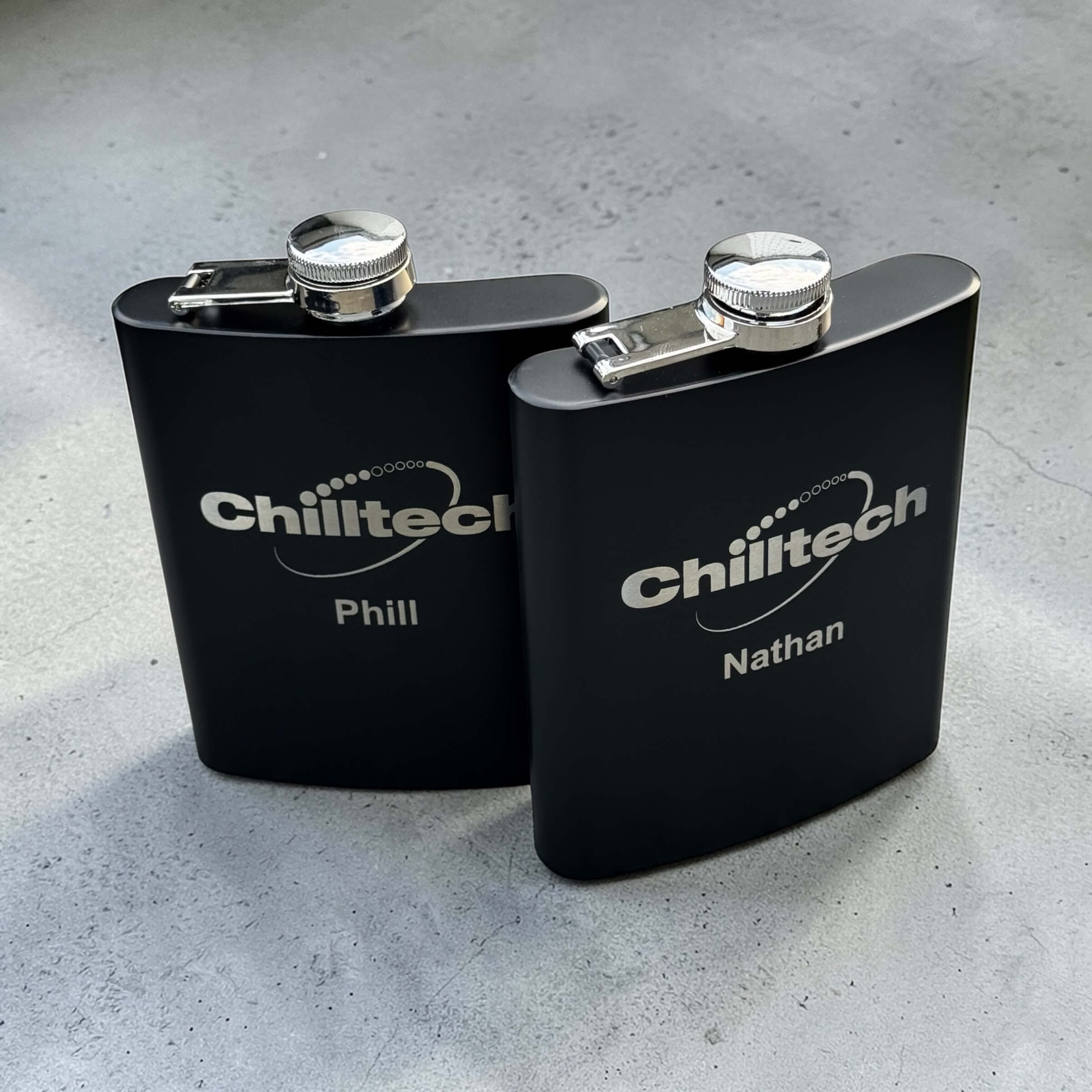 Two custom engraved black stainless steel hip flasks with corporate logo and personalised names, branded bulk corporate gifts