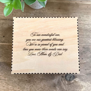 Wooden box for flask with engraved message on a wooden surface with a plant in the corner.