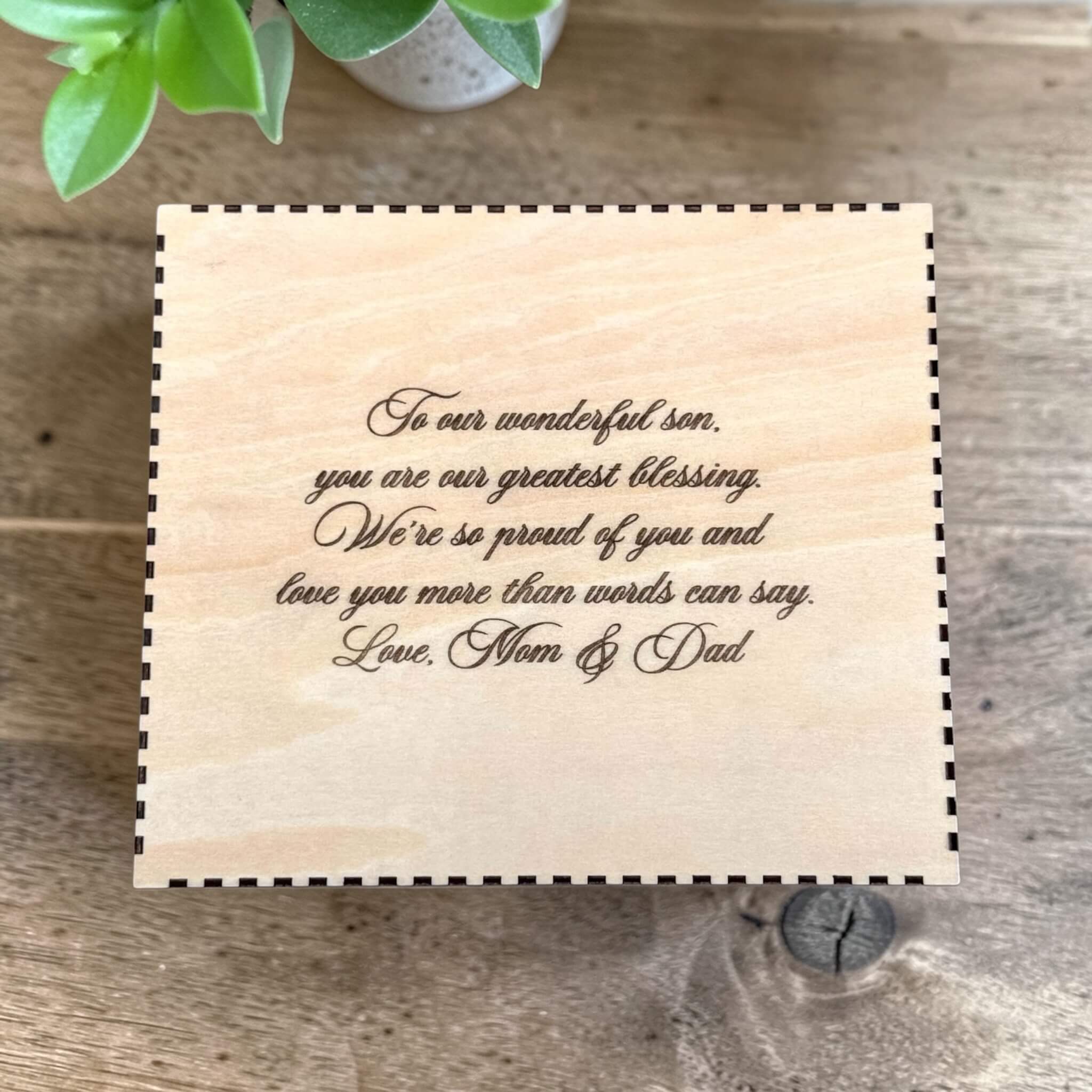 Wooden box for flask with engraved message on a wooden surface with a plant in the corner.