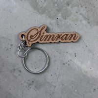 Wooden keychain with 'Simran' engraved on a concrete surface