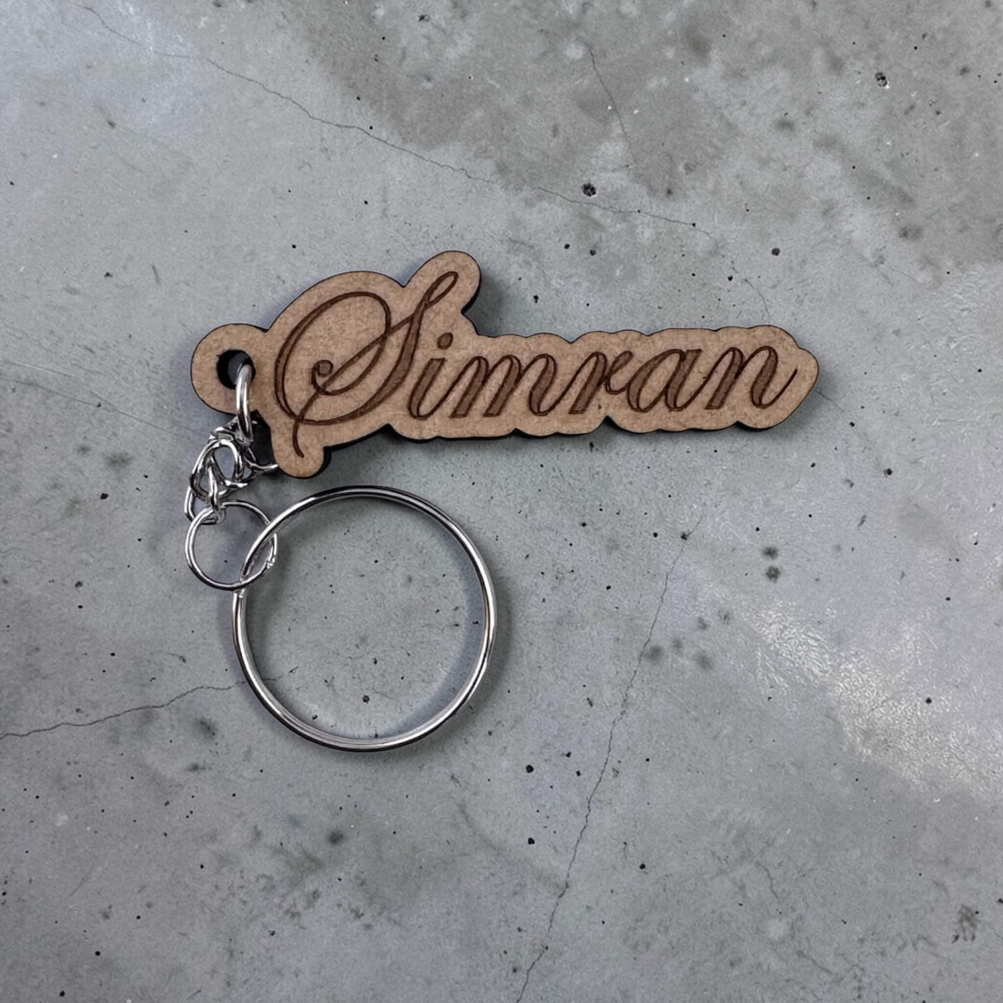 Wooden keychain with 'Simran' engraved on a concrete surface