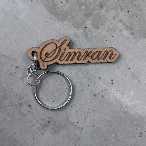 Wooden keychain with 'Simran' engraved on a concrete surface