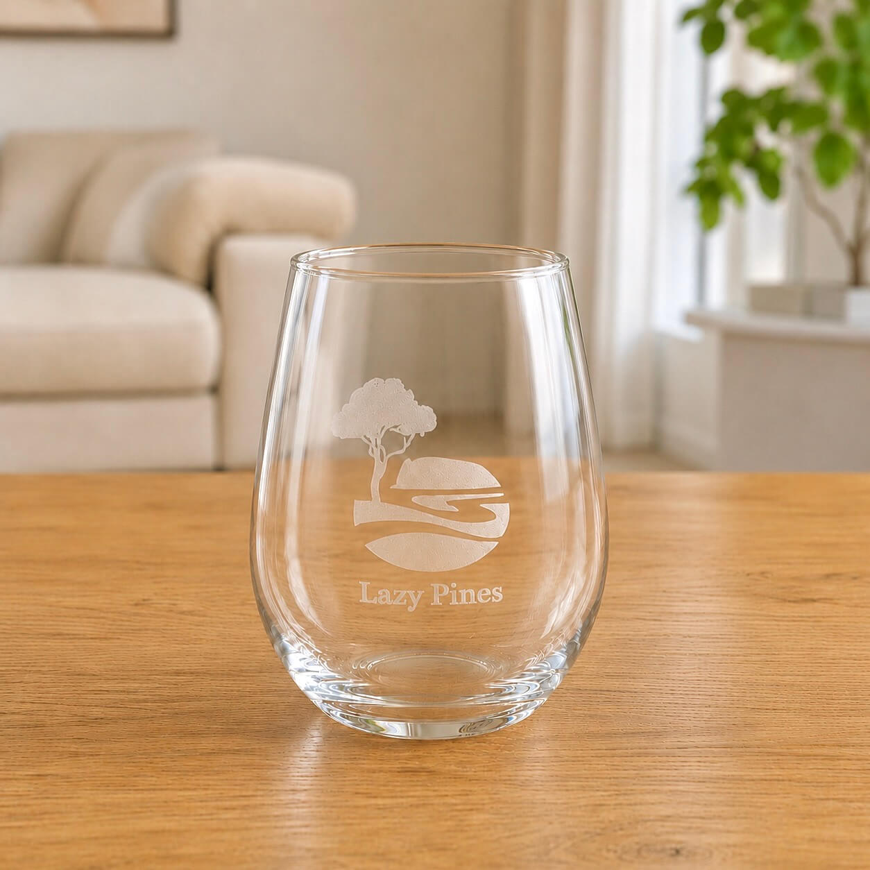 Close-up of clear stemless wine glass with etched Lazy Pines tree logo on wooden table